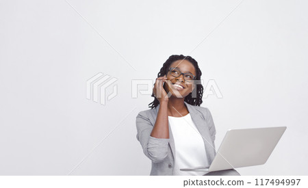 Cheerful black businesswoman talking on the phone and using a laptop, isolated on a white background, looking at copy space 117494997