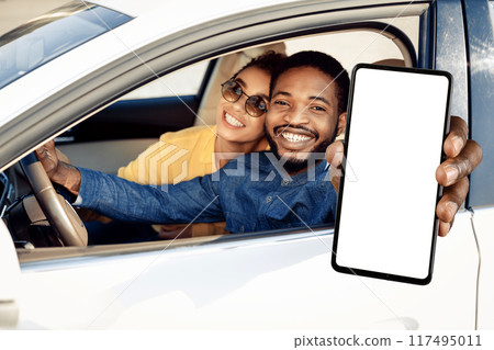A smiling young black man sits in the drivers seat of a car, holding a smartphone with a blank white screen out the window. A smiling young woman sits beside him. 117495011