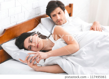 A woman is lying in bed, looking at her phone while a man, who is also in bed, watches her suspiciously. This image can be used to illustrate infidelity, mistrust, or relationship problems. A woman is lying in bed, looking at her phone while a man, who is also in bed, watches her suspiciously. This image can be used to illustrate infidelity, mistrust, or relationship problems. 117495061