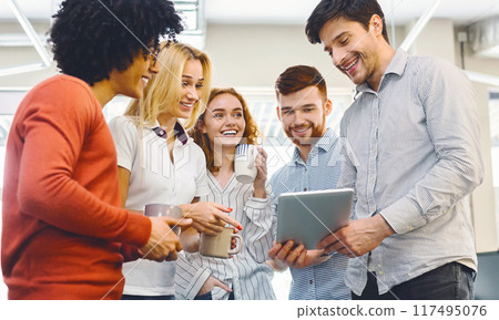 A diverse group of individuals standing side by side, looking attentively at a tablet device in the center. They appear engaged, possibly discussing or collaborating on the digital content displayed. A diverse group of individuals standing side by side, looking attentively at a tablet device in the center. They appear engaged, possibly discussing or collaborating on the digital content displayed. 117495076
