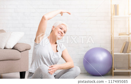 Positive active mature woman warming up, doing stretching exercises at home, free space Positive active mature woman warming up, doing stretching exercises at home, free space 117495085