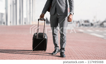 Unrecognizable black businessman walking with rolling suitcase outside a modern building, ready for a business trip or travel. Unrecognizable black businessman walking with rolling suitcase outside a modern building, ready for a business trip or travel. 117495088