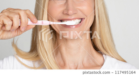 A close-up image of a woman with blonde hair smiling as she brushes her teeth with a pink toothbrush. The woman appears to be in her late 40s or early 50s, and her white teeth are visible A close-up image of a woman with blonde hair smiling as she brushes her teeth with a pink toothbrush. The woman appears to be in her late 40s or early 50s, and her white teeth are visible 117495164
