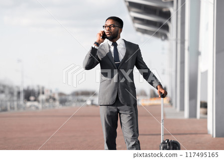 An executive black man talking on his phone with a suitcase at the airport entrance. The atmosphere is vibrant and professional, reflecting the fast-paced world of business travel. 117495165