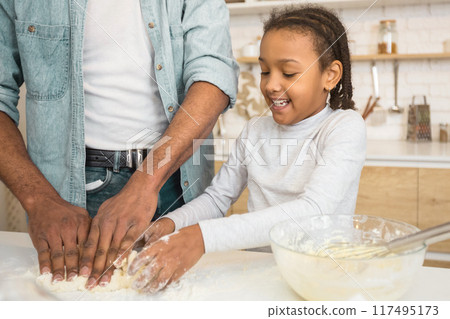 In a cozy kitchen, an african american father guides his daughter through the steps of kneading dough, symbolizing family time and fatherhood In a cozy kitchen, an african american father guides his daughter through the steps of kneading dough, symbolizing family time and fatherhood 117495173