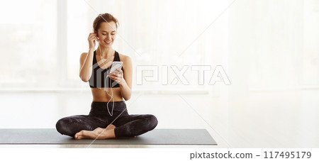 A young woman is seated in a cross-legged position on a yoga mat, wearing workout attire. She is listening to music through earphones, smiling as she enjoys her morning yoga session 117495179