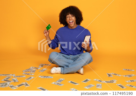 A young man with an curly sits on the floor with his legs crossed, holding a credit card in one hand and a smartphone in the other. The floor is covered in scattered cash. 117495190