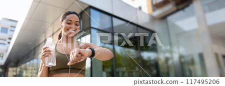 Young black woman looking at smartwatch, tracking fitness activity, listening to music in earphones, drinking water on training outdoors. Fit African American lady checking heart rate, copy space Young black woman looking at smartwatch, tracking fitness activity, listening to music in earphones, drinking water on training outdoors. Fit African American lady checking heart rate, copy space 117495206