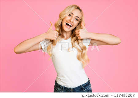 This image shows a happy blonde woman wearing a white t-shirt and blue jeans. She is giving a thumbs up with both hands and smiling. The background is a vibrant pink. This image shows a happy blonde woman wearing a white t-shirt and blue jeans. She is giving a thumbs up with both hands and smiling. The background is a vibrant pink. 117495207