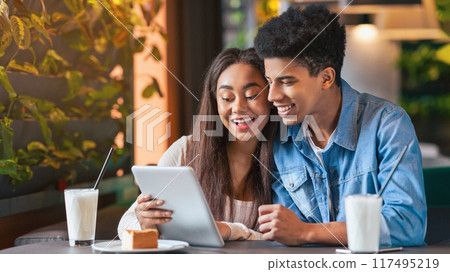 A young black guy and girl are sitting at a cafe table, engrossed in a tablet computer. They appear to be discussing something while looking at the screen, occasionally tapping and swiping. 117495219