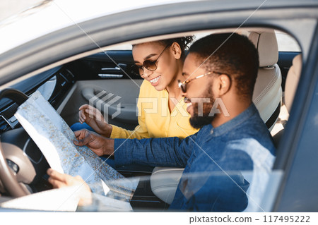African American couple in a car looks at a paper map to plan their road trip. The man, wearing a denim shirt and sunglasses, points at the map, while the woman looks at the map with a smile 117495222