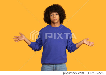 A young man with an curly hairstyle is standing in front of a yellow background. He is wearing a blue sweater and jeans. He is gesturing with both hands, palms facing outwards A young man with an curly hairstyle is standing in front of a yellow background. He is wearing a blue sweater and jeans. He is gesturing with both hands, palms facing outwards 117495226