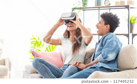 African American teen girl wearing a virtual reality headset smiles excitedly as she experiences a virtual reality game or simulation. She is sitting on a couch with guy companion who is smiling 117495232