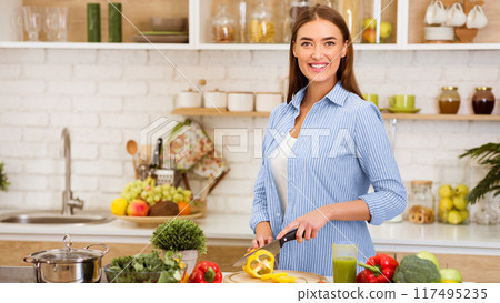 A young woman smiles as she chops a yellow bell pepper in a modern kitchen. 117495235