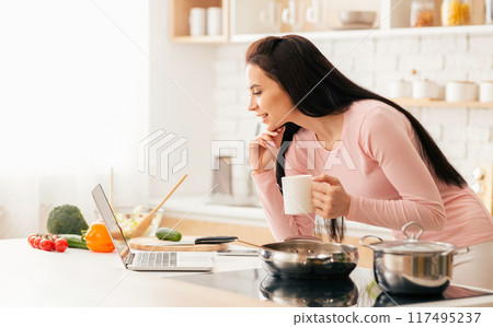 The woman is in a bright, modern kitchen looking at her laptop while holding a mug. She is preparing breakfast, surrounded by fresh vegetables and cooking utensils. The woman is in a bright, modern kitchen looking at her laptop while holding a mug. She is preparing breakfast, surrounded by fresh vegetables and cooking utensils. 117495237