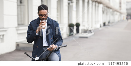Black man professional is riding a bicycle to the office, carrying coffee and checking phone. The scene is modern and busy, capturing the essence of multitasking and an active start to the day. Black man professional is riding a bicycle to the office, carrying coffee and checking phone. The scene is modern and busy, capturing the essence of multitasking and an active start to the day. 117495256