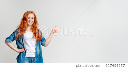 A cheerful red-haired woman stands against a plain background, one hand on her hip and the other extended as she points to the vacant area beside her, copy space A cheerful red-haired woman stands against a plain background, one hand on her hip and the other extended as she points to the vacant area beside her, copy space 117495257