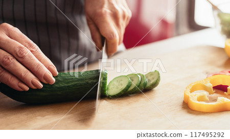 A woman hand slices a green cucumber on a wooden cutting board in a kitchen, with already sliced cucumbers and a sliced bell pepper in the background, cropped 117495292