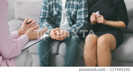 A therapist sits on a grey couch with a couple in front of them. The therapist is taking notes and listening to the couple. Therapist taking notes, possibly about the couples experiences or concerns. A therapist sits on a grey couch with a couple in front of them. The therapist is taking notes and listening to the couple. Therapist taking notes, possibly about the couples experiences or concerns. 117495293