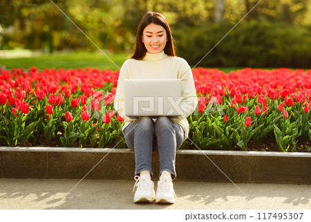 Asian young woman is sitting on a park ledge surrounded by blooming red tulips, using a laptop on a sunny spring day. She appears focused and happy in the vibrant garden setting. Asian young woman is sitting on a park ledge surrounded by blooming red tulips, using a laptop on a sunny spring day. She appears focused and happy in the vibrant garden setting. 117495307