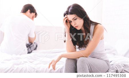 The image shows a woman sitting on a bed with her head in her hand. She is looking down and appears to be sad. A man is sitting behind her on the bed, with his head down The image shows a woman sitting on a bed with her head in her hand. She is looking down and appears to be sad. A man is sitting behind her on the bed, with his head down 117495308