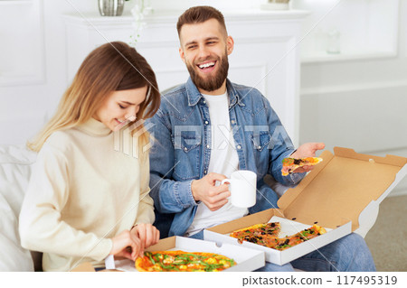 A man and woman are seated on a couch, enjoying pizza together. 117495319