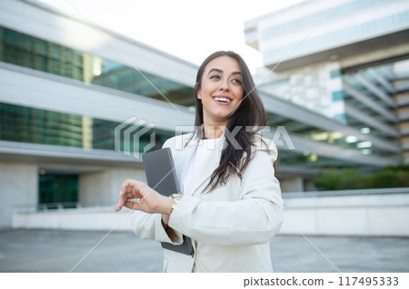 A young woman in a white blazer smiles while looking at her watch. She is holding a laptop and is standing outside a modern office building. The sky is bright and sunny. A young woman in a white blazer smiles while looking at her watch. She is holding a laptop and is standing outside a modern office building. The sky is bright and sunny. 117495333