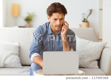 A man sits on a couch, using his laptop and talking on his phone. He looks concentrated and focused. A man sits on a couch, using his laptop and talking on his phone. He looks concentrated and focused. 117495369