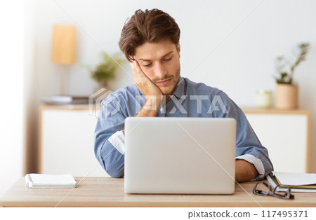 A tired man sits at his desk, looking weary as he rests his head on his hand while working on his laptop. A tired man sits at his desk, looking weary as he rests his head on his hand while working on his laptop. 117495371