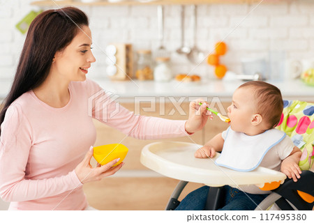 A baby boy is sitting in a high chair while his mother feeds him in the kitchen. The scene is warm and nurturing, with a cozy kitchen background and a loving interaction. A baby boy is sitting in a high chair while his mother feeds him in the kitchen. The scene is warm and nurturing, with a cozy kitchen background and a loving interaction. 117495380