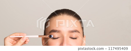 A woman is using a brush to apply makeup on her eyes. She is carefully brushing each eyelids to achieve a well-defined and neat look. 117495409