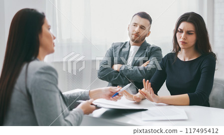 A couple is sitting at a table in an office, meeting with a lawyer. The woman is gesturing with her hand, while the man has his arms crossed. 117495412