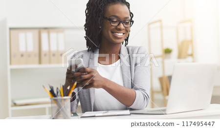 African American businesswoman smiling while using smartphone at her office desk, engaging in mobile communication. African American businesswoman smiling while using smartphone at her office desk, engaging in mobile communication. 117495447