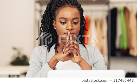 Pensive African American woman is sitting in front of a laptop computer, typing on the keyboard with a focused expression. The screen displays graphs and data, indicating work or research being done. 117495448
