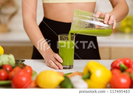 A woman pours a green smoothie from a blender into a glass on a kitchen counter surrounded by fresh produce. A woman pours a green smoothie from a blender into a glass on a kitchen counter surrounded by fresh produce. 117495483