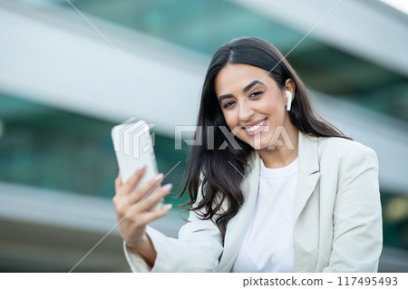 A young woman with dark hair, wearing a white t-shirt and a light blazer, stands in an urban setting. She smiles at the camera as she holds up her smartphone and appears to be using it. 117495493