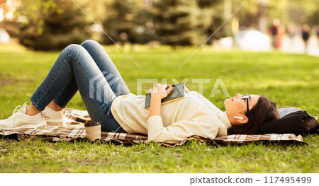 Asian young woman wearing casual clothes is lying on a blanket, holding a book, and listening to music with wireless earbuds. She is surrounded by lush green grass and trees on a sunny day 117495499