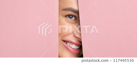 Cheerful woman with her hair in a bun and wearing a white shirt is peeking out of a hole in a vibrant pink wall. Cheerful woman with her hair in a bun and wearing a white shirt is peeking out of a hole in a vibrant pink wall. 117495530
