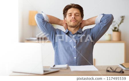 A man sits at his desk with a laptop, eyes closed and hands behind his head, taking a moment to relax. A man sits at his desk with a laptop, eyes closed and hands behind his head, taking a moment to relax. 117495539