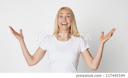 A woman with blonde hair smiles excitedly and opens her arms wide, as if she is surprised and happy. She is wearing a white t-shirt and is standing against a white background A woman with blonde hair smiles excitedly and opens her arms wide, as if she is surprised and happy. She is wearing a white t-shirt and is standing against a white background 117495594