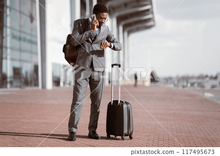 African American businessman on the phone with rolling suitcase outside a modern building, checking time, preparing for a business trip. 117495673