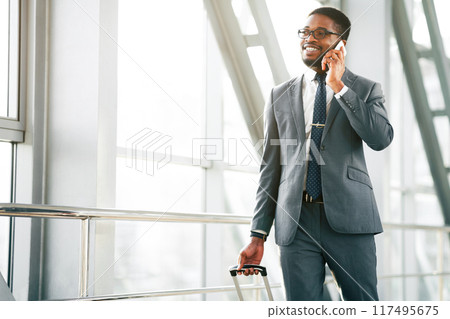 African American professional traveler talking on his phone outside a sleek airport, holding his luggage. The mood is focused and efficient, capturing the essence of a business trip, copy space 117495675