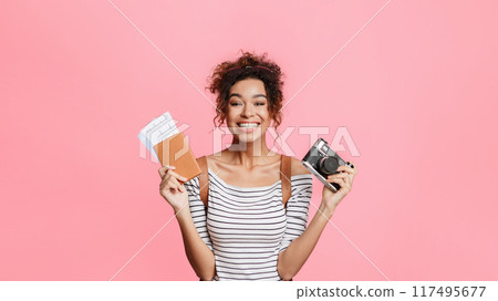 African-american woman holding camera and passport with tickets, pink background African-american woman holding camera and passport with tickets, pink background 117495677