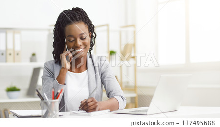 Delighted young African American woman in professional outfit, using a laptop at her office desk, chatting on the phone. 117495688