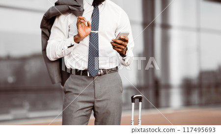 Cropped of black businessman in a suit checking his phone while standing with a rolling suitcase outside a modern building, copy space Cropped of black businessman in a suit checking his phone while standing with a rolling suitcase outside a modern building, copy space 117495693