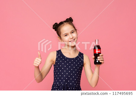 A young girl with long brown hair and wearing a pink shirt is holding a bottle of soda in one hand. She is smiling and giving a thumbs up gesture with her other hand. 117495695