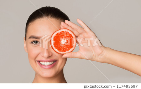A woman is shown holding a slice of grapefruit over her face. The fruit is positioned in front of her eyes, nose, and mouth. She appears to be examining the grapefruit closely. A woman is shown holding a slice of grapefruit over her face. The fruit is positioned in front of her eyes, nose, and mouth. She appears to be examining the grapefruit closely. 117495697