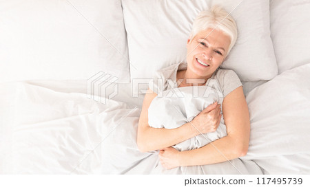 Cheerful senior woman relaxing in bed, morning awakening, top view with free space Cheerful senior woman relaxing in bed, morning awakening, top view with free space 117495739