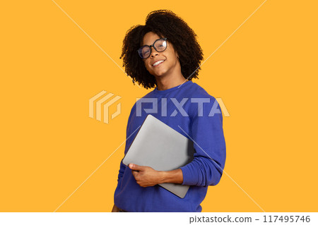 This is a photo of a smiling young man with curly black hair wearing glasses and a blue sweatshirt. He is holding a silver laptop in front of him. He is standing in front of bright yellow background. This is a photo of a smiling young man with curly black hair wearing glasses and a blue sweatshirt. He is holding a silver laptop in front of him. He is standing in front of bright yellow background. 117495746