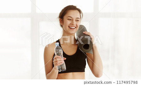 A woman standing while holding a water bottle in one hand and a yoga mat in the other hand. Healthy lifestyle concept A woman standing while holding a water bottle in one hand and a yoga mat in the other hand. Healthy lifestyle concept 117495798
